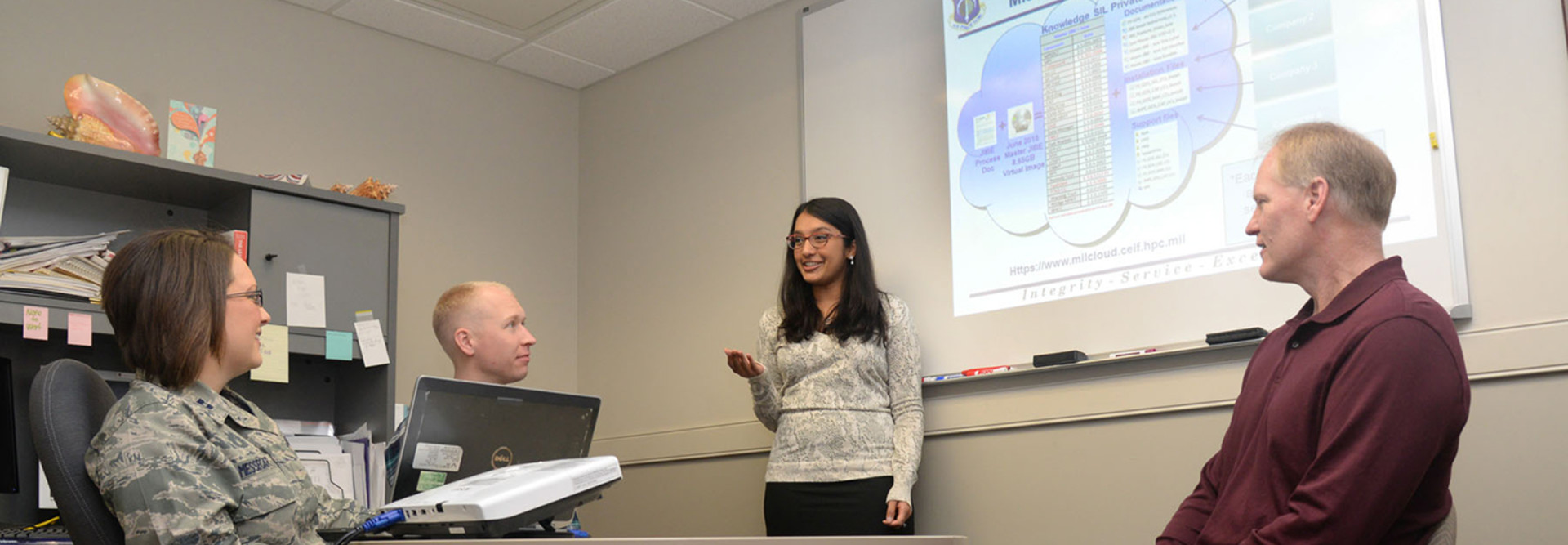 Ammu Irivinti, MITRE senior systems engineer, standing, speaks with, from left to right, Capt. Devon Messecar, System Engineering Integration Contract II program manager, Steve Harrison, MITRE Advanced Development lead, and Dave Parker, deputy PM, at Hanscom Air Force Base, Mass., on Nov. 13, 2015, regarding the use of Hanscom MilCloud to improve mission planning.