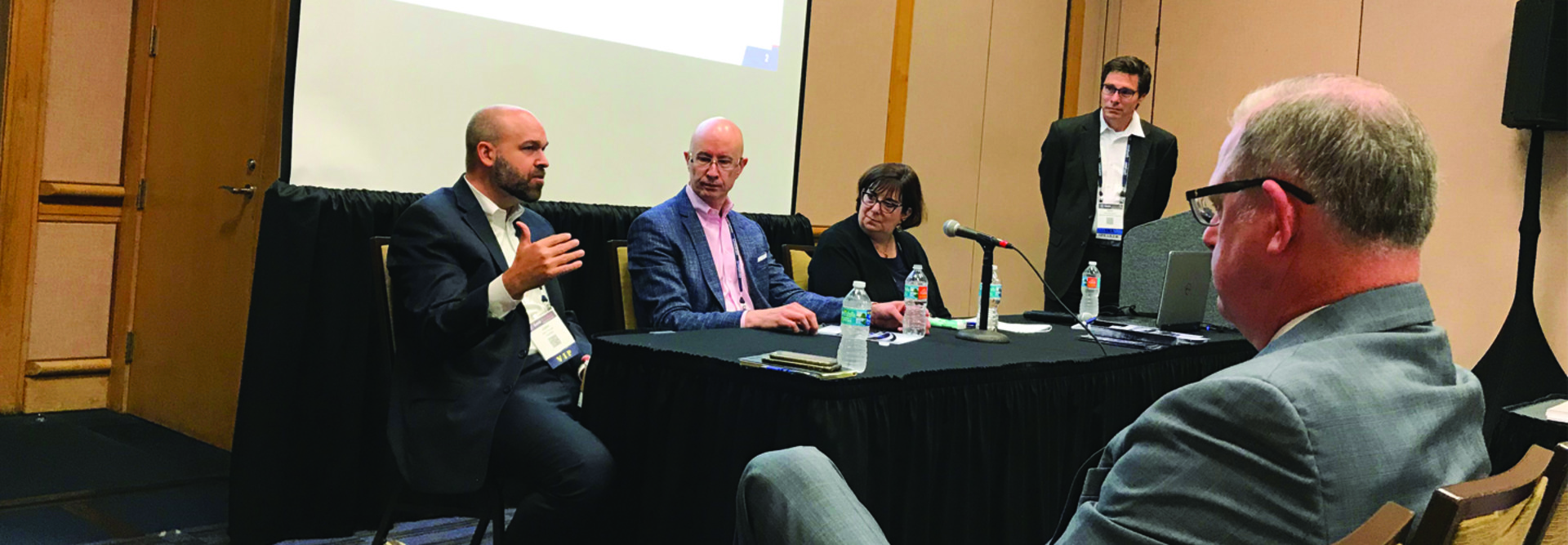 Two bald men sitting at a table next to a woman, one man standing at a podium, one man sitting and listening