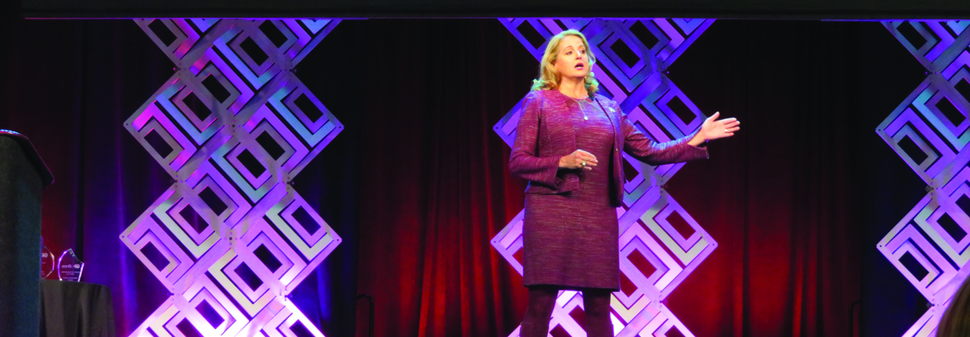 blond woman in cranberry colored suit gesturing on a stage