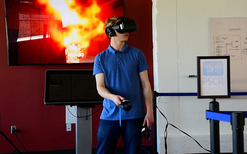 A NIST researcher demonstrates the use of a virtual reality headset and controllers in the agency’s virtual office environment, in which first responders train to search for a body in a fire.