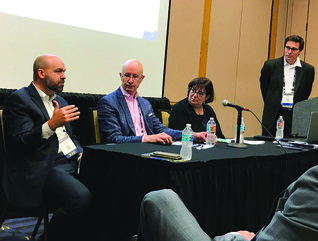 Two bald men sitting at a table next to a woman, one man standing at a podium, one man sitting and listening