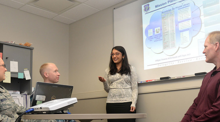 Ammu Irivinti, MITRE senior systems engineer, standing, speaks with, from left to right, Capt. Devon Messecar, System Engineering Integration Contract II program manager, Steve Harrison, MITRE Advanced Development lead, and Dave Parker, deputy PM, at Hanscom Air Force Base, Mass., on Nov. 13, 2015, regarding the use of Hanscom MilCloud to improve mission planning.