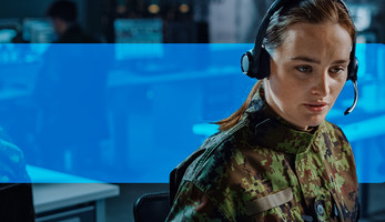 Woman with headset on, in a room with other people working on computers.