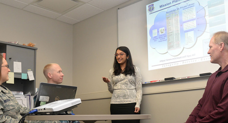 Ammu Irivinti, MITRE senior systems engineer, standing, speaks with, from left to right, Capt. Devon Messecar, System Engineering Integration Contract II program manager, Steve Harrison, MITRE Advanced Development lead, and Dave Parker, deputy PM, at Hanscom Air Force Base, Mass., on Nov. 13, 2015, regarding the use of Hanscom MilCloud to improve mission planning.